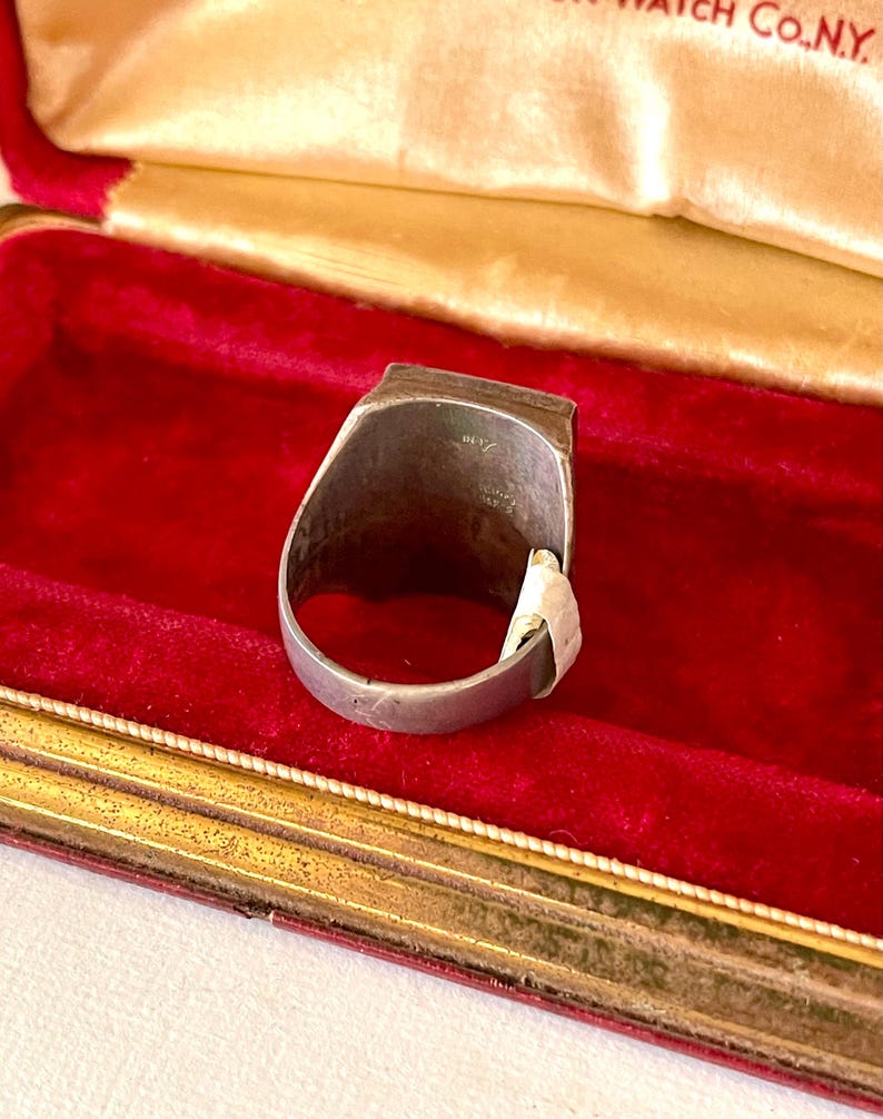 K&ouml;nnte beinhalten: Ein silberfarbener Ring mit rechteckiger Oberseite, pr&auml;sentiert in einer roten Samt-Schmuckschatulle. Der Ring hat ein schlichtes Band und ein leicht abgenutztes Aussehen. Die Schatulle hat einen goldfarbenen Rand und ein cremefarbenes Satinfutter.