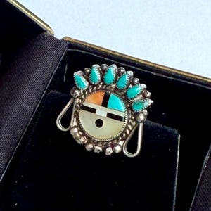 May include: A silver ring featuring a circular inlay face with a geometric design in turquoise, orange, and white stone, accented by a black horizontal bar and dot. The setting is surrounded by a row of small turquoise teardrop stones.