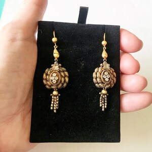 May include: A pair of antique gold-toned earrings with intricate detailing. Each earring features a central oval design with clear stones, surrounded by a textured border and a dangling tassel. The earrings are displayed on a black velvet background.