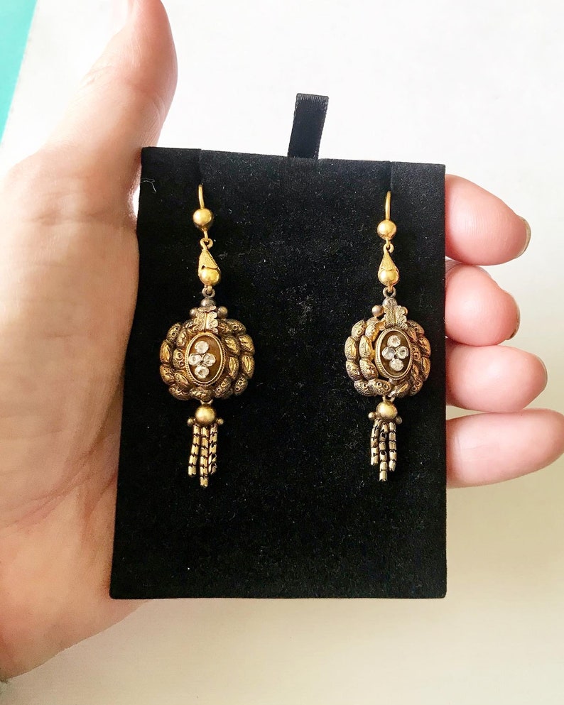 May include: A pair of antique gold-toned earrings with intricate detailing. Each earring features a central oval design with clear stones, surrounded by a textured border and a dangling tassel. The earrings are displayed on a black velvet background.