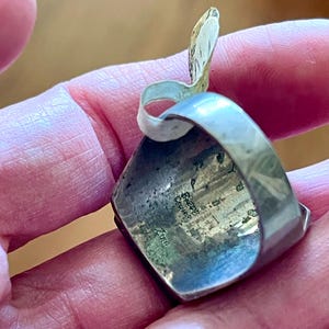 K&ouml;nnte beinhalten: Ein silberner Ring mit einem quadratischen, dunkel gef&auml;rbten Stein. Der Ring hat ein einzigartiges Design mit einem gebogenen, blattartigen Element. Die Oberfl&auml;che des Rings weist Gebrauchsspuren auf, was darauf hindeutet, dass er Vintage oder antik ist. Der Ring wird in einer Hand gehalten.