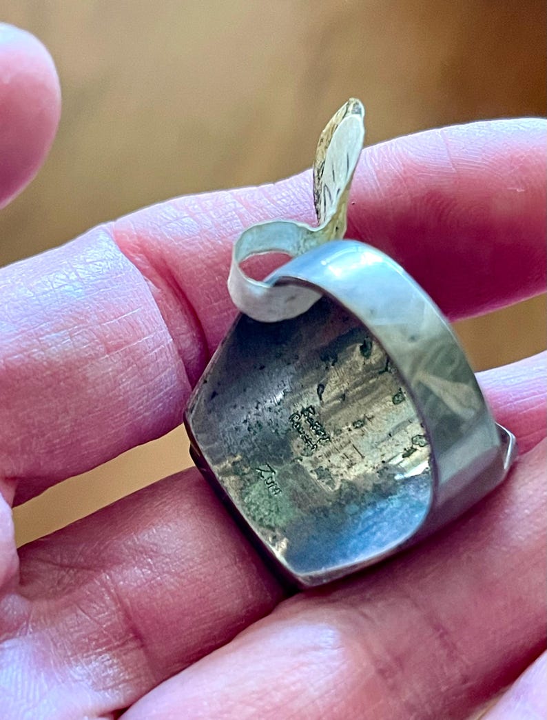 K&ouml;nnte beinhalten: Ein silberner Ring mit einem quadratischen, dunkel gef&auml;rbten Stein. Der Ring hat ein einzigartiges Design mit einem gebogenen, blattartigen Element. Die Oberfl&auml;che des Rings weist Gebrauchsspuren auf, was darauf hindeutet, dass er Vintage oder antik ist. Der Ring wird in einer Hand gehalten.