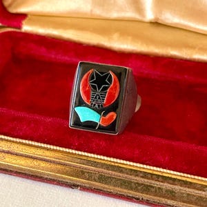 K&ouml;nnte beinhalten: Ein silberner Ring mit einem schwarzen quadratischen Gesicht mit einem Stern-, Halbmond- und Flaggen-Design in Rot, T&uuml;rkis und Schwarz. Der Ring wird in einer rot samtgef&uuml;tterten Schatulle mit goldenem Rand pr&auml;sentiert.
