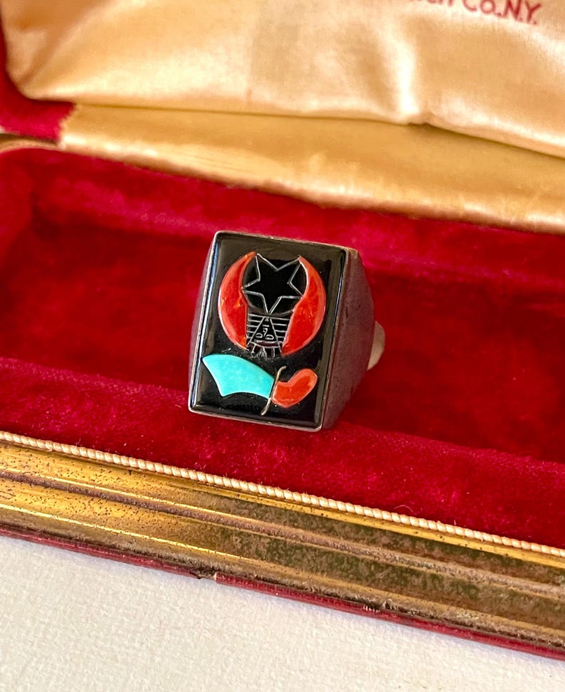 K&ouml;nnte beinhalten: Ein silberner Ring mit einem schwarzen quadratischen Gesicht mit einem Stern-, Halbmond- und Flaggen-Design in Rot, T&uuml;rkis und Schwarz. Der Ring wird in einer rot samtgef&uuml;tterten Schatulle mit goldenem Rand pr&auml;sentiert.