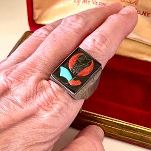 K&ouml;nnte beinhalten: Rechteckiger Silberring mit schwarzem Inlay, das einen Stern, eine Mondsichel und andere Formen in Rot, T&uuml;rkis und Wei&szlig; zeigt. Der Ring befindet sich an einem Finger, mit einer roten Samtbox im Hintergrund. Die Box tr&auml;gt die Aufschrift "MT VERNON Made in U.S.A."