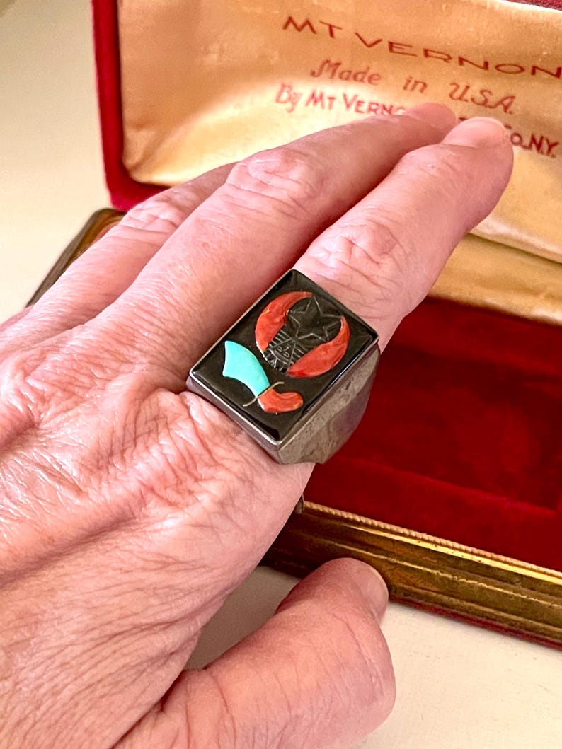 K&ouml;nnte beinhalten: Rechteckiger Silberring mit schwarzem Inlay, das einen Stern, eine Mondsichel und andere Formen in Rot, T&uuml;rkis und Wei&szlig; zeigt. Der Ring befindet sich an einem Finger, mit einer roten Samtbox im Hintergrund. Die Box tr&auml;gt die Aufschrift "MT VERNON Made in U.S.A."