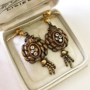 May include: A pair of antique gold-toned earrings with a floral design and clear gemstones. The earrings are in a white box with the text "F. Lameroux, Lisieux" on the lid.