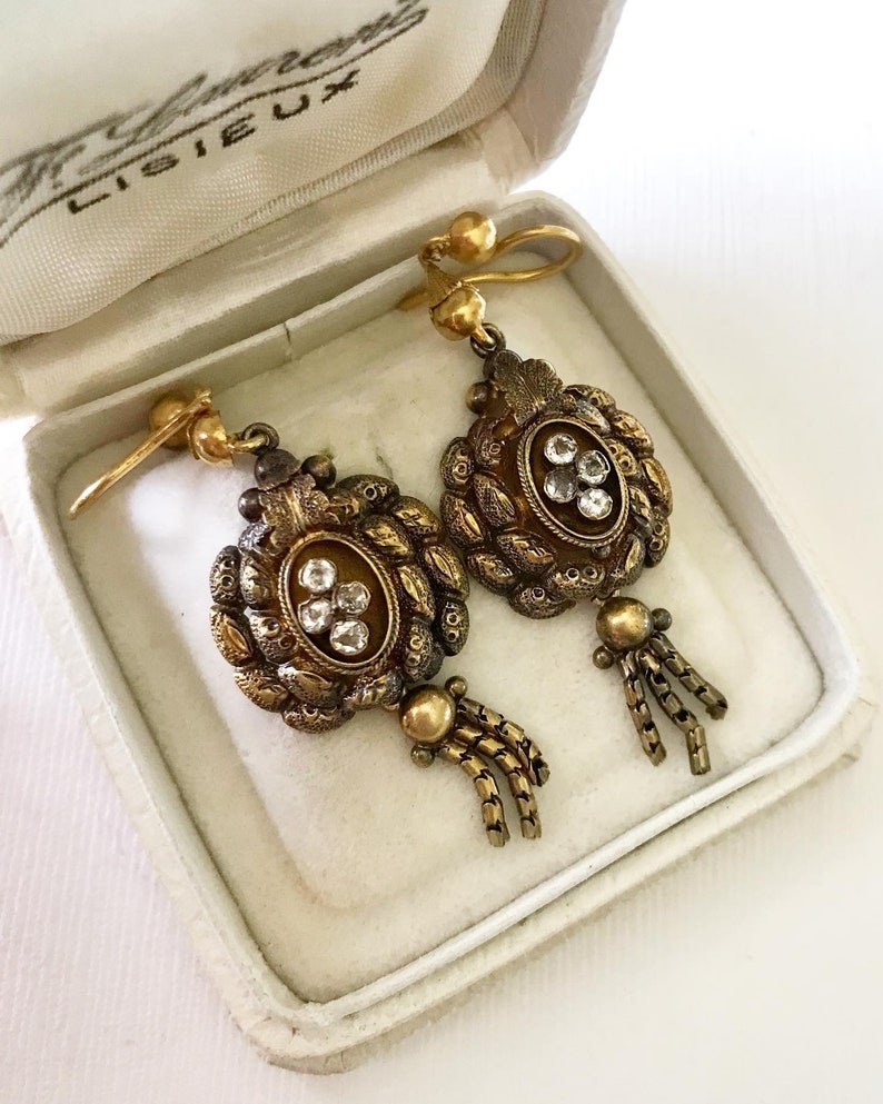 May include: A pair of antique gold-toned earrings with a floral design and clear gemstones. The earrings are in a white box with the text "F. Lameroux, Lisieux" on the lid.