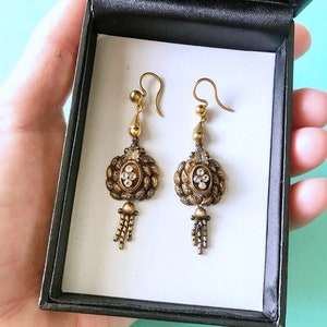 May include: A pair of antique gold-toned earrings with a drop design. Each earring features an oval centerpiece with clear stones, surrounded by ornate detailing and a small tassel. The earrings are displayed in a black box with a white interior.