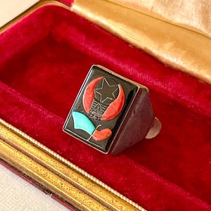 K&ouml;nnte beinhalten: Ein silberner Ring mit rechteckiger schwarzer Oberfl&auml;che, die einen Stern, eine Mondsichel und ein geometrisches Design in Rot, T&uuml;rkis und Wei&szlig; zeigt. Der Ring liegt in einer roten Samt-Schmuckschatulle.