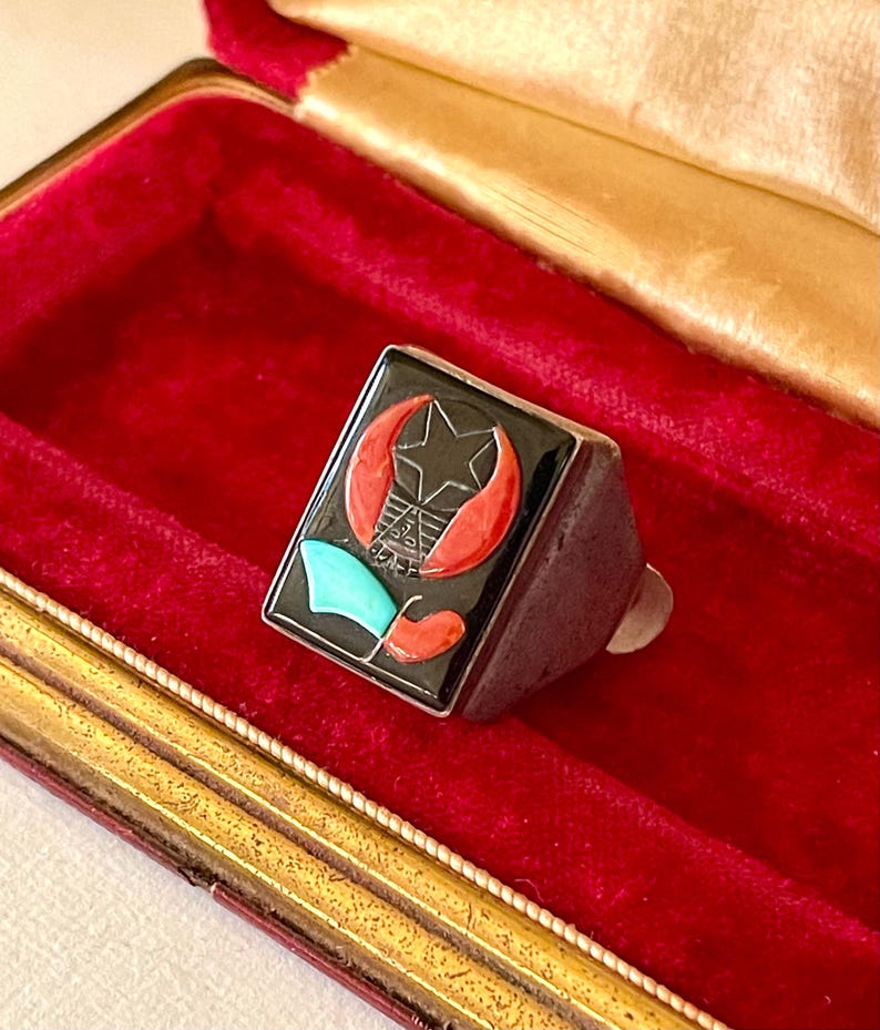 K&ouml;nnte beinhalten: Ein silberner Ring mit rechteckiger schwarzer Oberfl&auml;che, die einen Stern, eine Mondsichel und ein geometrisches Design in Rot, T&uuml;rkis und Wei&szlig; zeigt. Der Ring liegt in einer roten Samt-Schmuckschatulle.