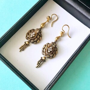 May include: A pair of antique gold-toned earrings with intricate detailing. Each earring features a central oval design with clear stones, surrounded by ornate metalwork. Below the oval is a small tassel. The earrings are displayed in a black box with a white interior.