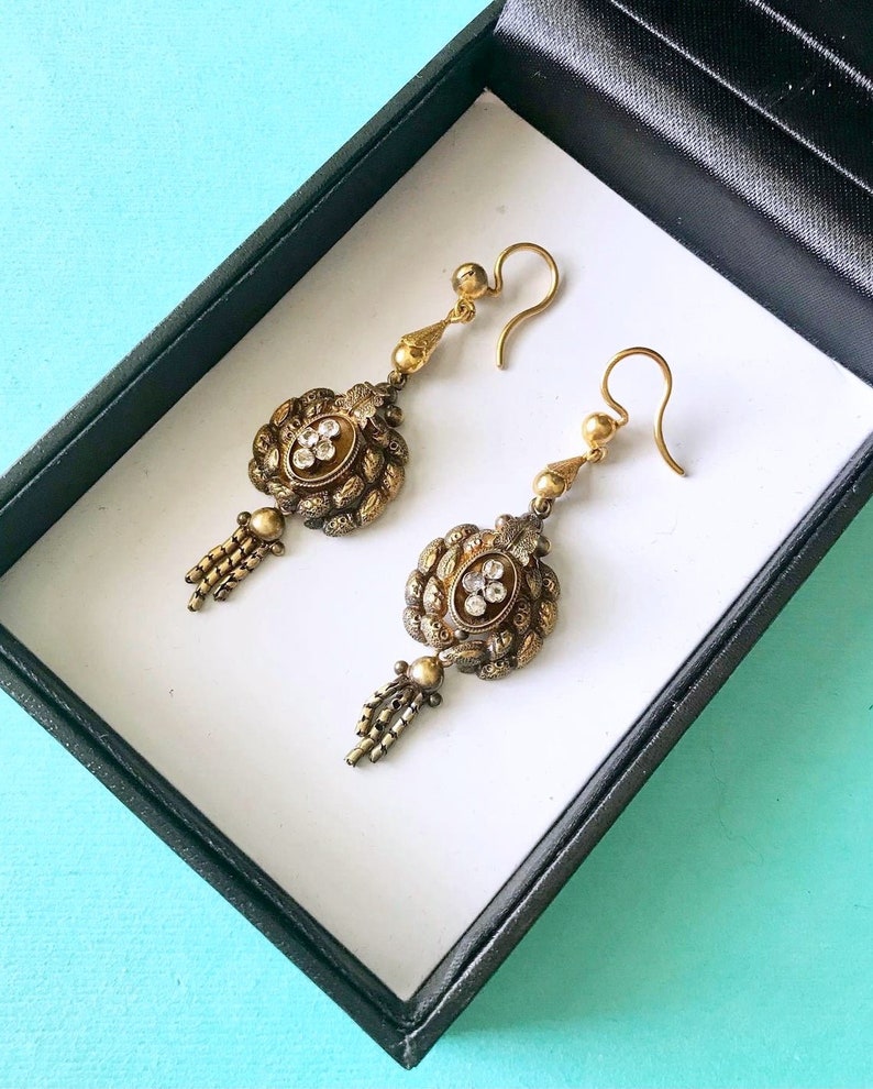May include: A pair of antique gold-toned earrings with intricate detailing. Each earring features a central oval design with clear stones, surrounded by ornate metalwork. Below the oval is a small tassel. The earrings are displayed in a black box with a white interior.