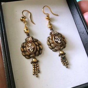May include: A pair of antique gold-toned earrings. Each earring features an oval-shaped centerpiece with clear stones and a tassel of small chains. The earrings are displayed in a black box with a white interior, suggesting they are for sale.