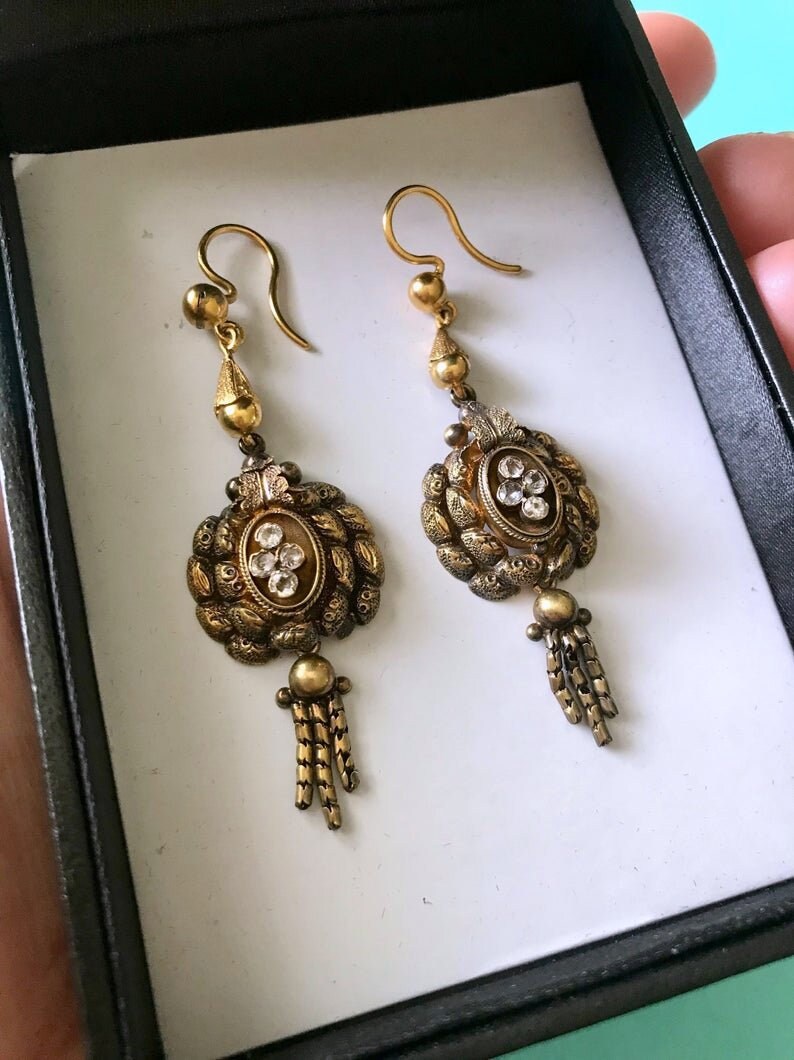May include: A pair of antique gold-toned earrings. Each earring features an oval-shaped centerpiece with clear stones and a tassel of small chains. The earrings are displayed in a black box with a white interior, suggesting they are for sale.