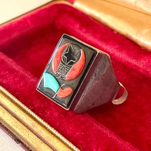 K&ouml;nnte beinhalten: Ein silberner Ring mit rechteckiger schwarzer Oberfl&auml;che, die einen Stern, einen Halbmond und andere geometrische Formen in Rot, T&uuml;rkis und Wei&szlig; zeigt. Der Ring liegt in einer roten Samtbox.