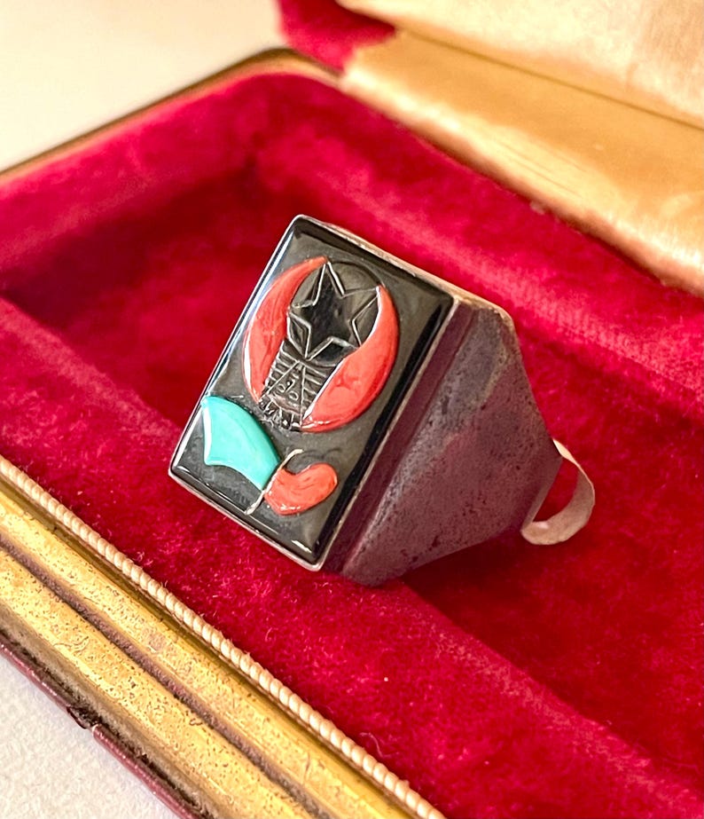 K&ouml;nnte beinhalten: Ein silberner Ring mit rechteckiger schwarzer Oberfl&auml;che, die einen Stern, einen Halbmond und andere geometrische Formen in Rot, T&uuml;rkis und Wei&szlig; zeigt. Der Ring liegt in einer roten Samtbox.