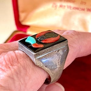 K&ouml;nnte beinhalten: Ein Silberring mit rechteckiger schwarzer Onyx-Oberseite. Die Oberseite zeigt einen roten Hummer, einen t&uuml;rkisfarbenen Akzent und ein rotes Paprikadesign. Der Ring wird am Finger getragen.