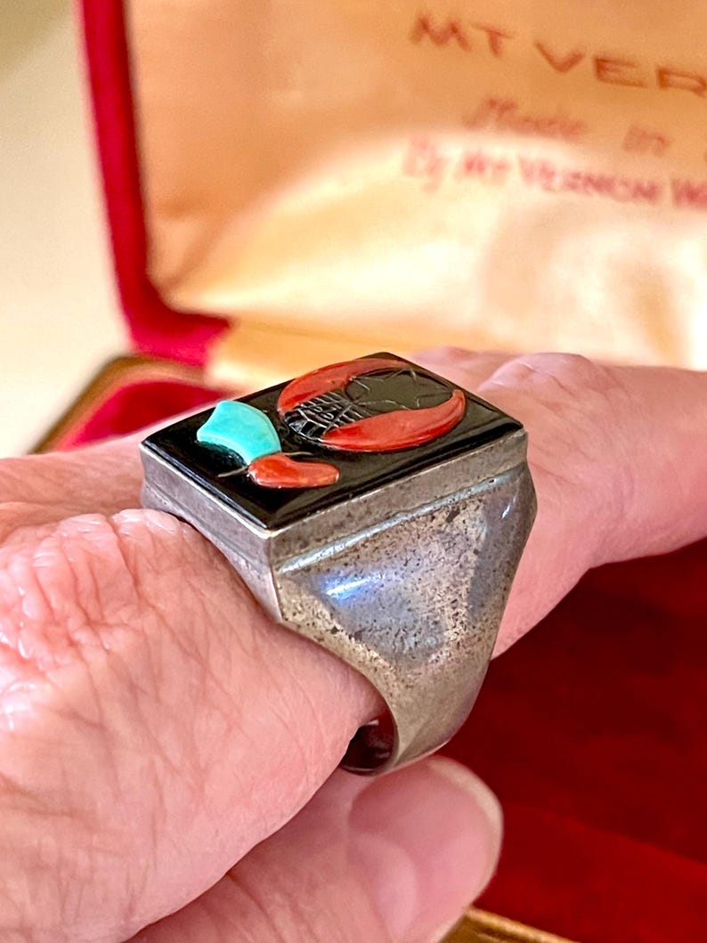 K&ouml;nnte beinhalten: Ein Silberring mit rechteckiger schwarzer Onyx-Oberseite. Die Oberseite zeigt einen roten Hummer, einen t&uuml;rkisfarbenen Akzent und ein rotes Paprikadesign. Der Ring wird am Finger getragen.