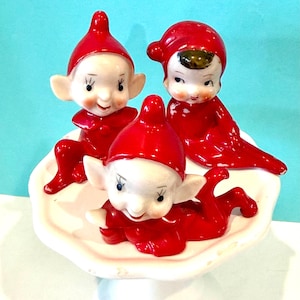 May include: Three ceramic elf figurines wearing bright red outfits and pointed hats. Two elves are seated upright with pointed ears, while the third elf is lying down. They are arranged on a white, scalloped-edge pedestal dish with a glossy finish.
