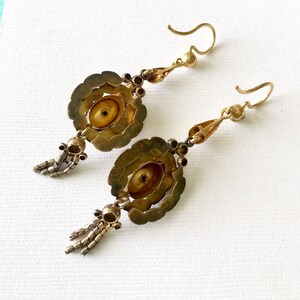 May include: A pair of antique gold-toned earrings. Each earring features a floral design with a central, circular element. Small, dangling chains and decorative beads add detail. The earrings have a hook closure.