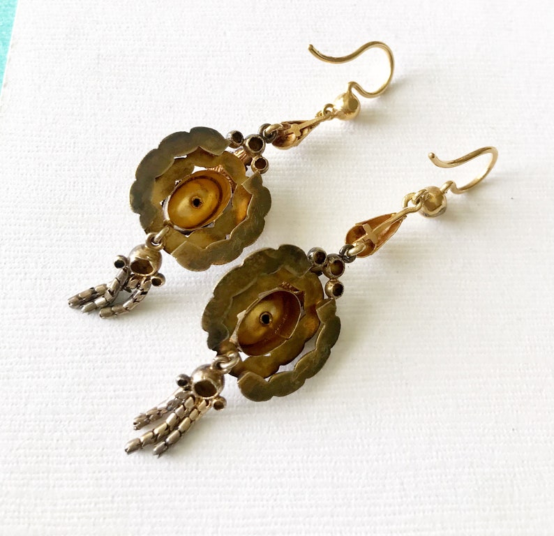 May include: A pair of antique gold-toned earrings. Each earring features a floral design with a central, circular element. Small, dangling chains and decorative beads add detail. The earrings have a hook closure.