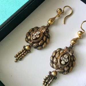 May include: A pair of antique gold-toned earrings. Each earring features an oval centerpiece with clear stones, surrounded by ornate detailing. Below the centerpiece is a small tassel. The earrings are displayed in a white box.