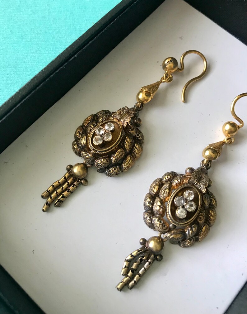 May include: A pair of antique gold-toned earrings. Each earring features an oval centerpiece with clear stones, surrounded by ornate detailing. Below the centerpiece is a small tassel. The earrings are displayed in a white box.