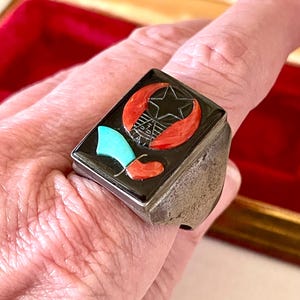 K&ouml;nnte beinhalten: Rechteckiger Silberring mit schwarzer Einlage, verziert mit einem Stern, einem Halbmond und anderen geometrischen Formen in Rot, T&uuml;rkis und Schwarz. Der Ring wird am Finger getragen und hat ein strukturiertes, gealtertes Aussehen.