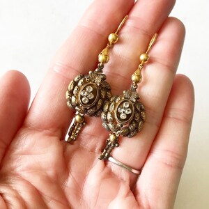May include: A pair of antique gold-toned earrings. Each earring features an oval centerpiece with clear, sparkling stones and intricate detailing. The earrings have a drop design with small chain accents and a hook closure.