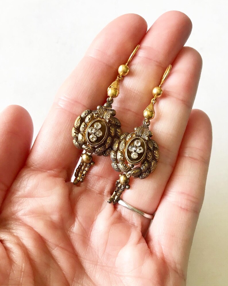 May include: A pair of antique gold-toned earrings. Each earring features an oval centerpiece with clear, sparkling stones and intricate detailing. The earrings have a drop design with small chain accents and a hook closure.