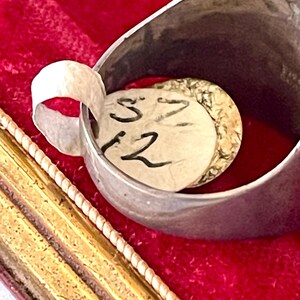 K&ouml;nnte beinhalten: Nahaufnahme eines Silberrings mit einem Papieranh&auml;nger im Inneren. Der Anh&auml;nger tr&auml;gt die handgeschriebenen Zahlen "S 12". Der Ring befindet sich vor einem roten Samthintergrund, mit einem goldfarbenen Rahmen im Vordergrund.