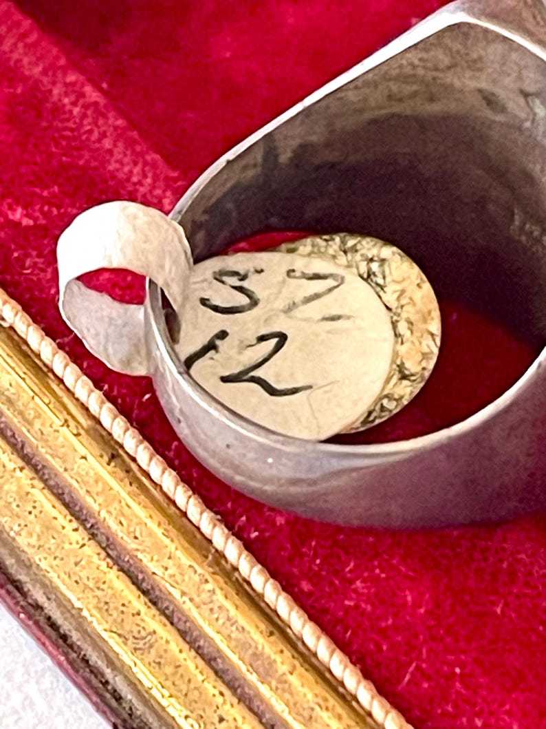 K&ouml;nnte beinhalten: Nahaufnahme eines Silberrings mit einem Papieranh&auml;nger im Inneren. Der Anh&auml;nger tr&auml;gt die handgeschriebenen Zahlen "S 12". Der Ring befindet sich vor einem roten Samthintergrund, mit einem goldfarbenen Rahmen im Vordergrund.