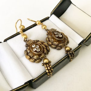 May include: A pair of antique gold-toned earrings with intricate floral and beaded designs. Each earring features a central oval setting with clear gemstones, surrounded by detailed metalwork and dangling accents. The earrings are displayed in a black jewelry box with a white interior.
