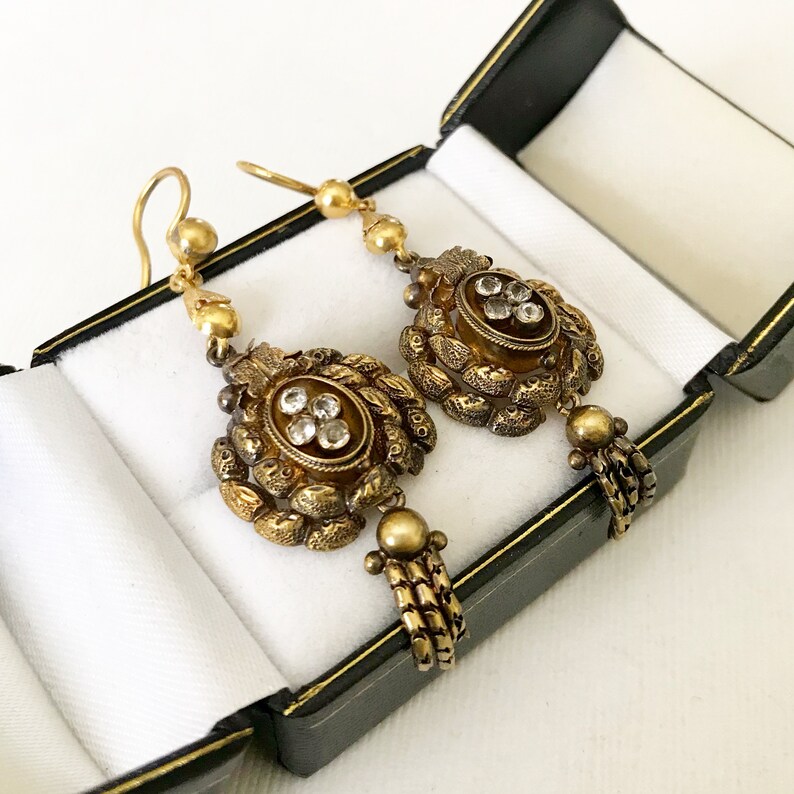 May include: A pair of antique gold-toned earrings with intricate floral and beaded designs. Each earring features a central oval setting with clear gemstones, surrounded by detailed metalwork and dangling accents. The earrings are displayed in a black jewelry box with a white interior.
