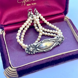 May include: A vintage pearl bracelet with three strands of small, off-white pearls. The bracelet features an ornate, silver-toned clasp with floral detailing. The bracelet is displayed inside a purple velvet jewelry box.