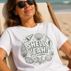 Puede incluir: Una camiseta blanca de manga corta con un estampado gráfico en verde oscuro de varias conchas marinas y una estrella de mar dispuestas en círculo. El texto central dice "SHELL YEAH!" en negrita con "IT'S SUMMER" impreso debajo.