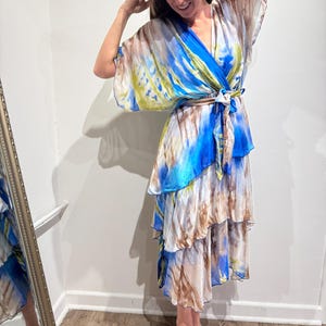 May include: A flowing, tie-dye dress in shades of blue, white, brown, and yellow. The dress features a wrap-style top, a belted waist, and a tiered skirt. The sleeves are wide and flowing.