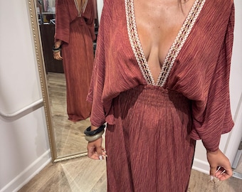Puede incluir: Un vestido largo color óxido con un escote en V profundo y mangas anchas. El escote está adornado con un diseño bordado en crema y dorado. El vestido tiene una cintura ceñida y una falda fluida. Se ve un collar de gargantilla dorado.