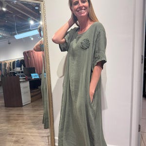 May include: An olive green maxi dress with short sleeves and a round neckline. The dress features a decorative floral embellishment on the chest and pockets. The woman is wearing brown sandals.