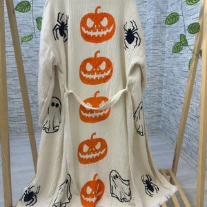 May include: Cream-colored Halloween robe featuring orange jack-o'-lanterns, black spiders, and white ghost designs. The robe has long sleeves and a matching belt. The robe is hanging on a wooden rack.
