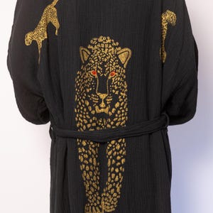 Op de afbeelding: Zwarte robe met een gouden luipaardprint. De robe heeft een strikceintuur en heeft een grote luipaardprint op de rug en kleinere luipaardprints op de mouwen.