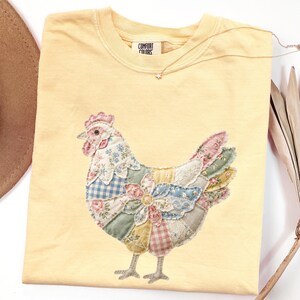 May include: A yellow Comfort Colors t-shirt featuring a patchwork chicken design. The chicken is made of various floral and patterned fabrics in shades of pink, blue, yellow, and green. A delicate star pendant necklace rests on the shirt.
