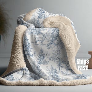May include: A cozy blanket with a forest scene in blue and white, featuring deer and trees. The blanket has a soft, cream-colored sherpa lining. The words "Ships Fast!" are visible on the blanket.