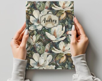 May include: A personalized notebook with a floral design featuring white magnolia flowers and green leaves. The cover has a brown label with the name "Audrey" and the letter "A" in a decorative font.