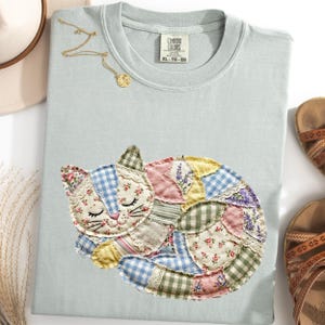 Könnte beinhalten: Hellblaues Comfort Colors T-Shirt mit einem Patchwork-Katzen-Design. Die Katze besteht aus verschiedenen floralen und gemusterten Stoffen in Pastellfarben. Eine goldene Halskette und Sandalen sind ebenfalls zu sehen.
