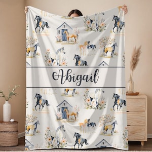 May include: A white blanket with a repeating pattern of watercolour horses, barns, and floral elements. The name "Abigail" is displayed in elegant script across the centre. A soft, cosy home decor item.