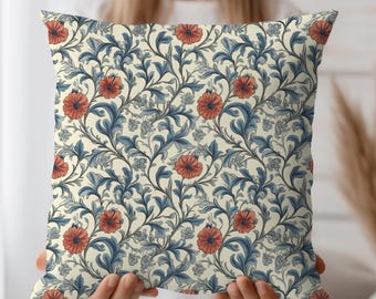May include: Square decorative pillow with a floral pattern. The pillow features a cream-colored background with a repeating design of red flowers and blue leaves. The pillow is held by a person.