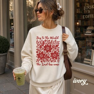May include: An ivory long-sleeve crewneck sweatshirt featuring a red graphic print of poinsettias, pine branches, and pinecones. The text "Joy to the World" is printed above the floral design, and "the Lord has come" is printed below the design.