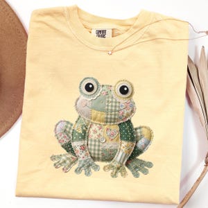 May include: Yellow Comfort Colors t-shirt featuring a patchwork frog design. The frog has large eyes and is made of various floral and patterned fabrics in shades of green, yellow, and pink. A delicate star necklace is visible.